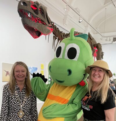 The Mayor standing in front of a giant dinosaur skeleton, alongside someone wearing a carton dinosaur costume and one of the museum staff