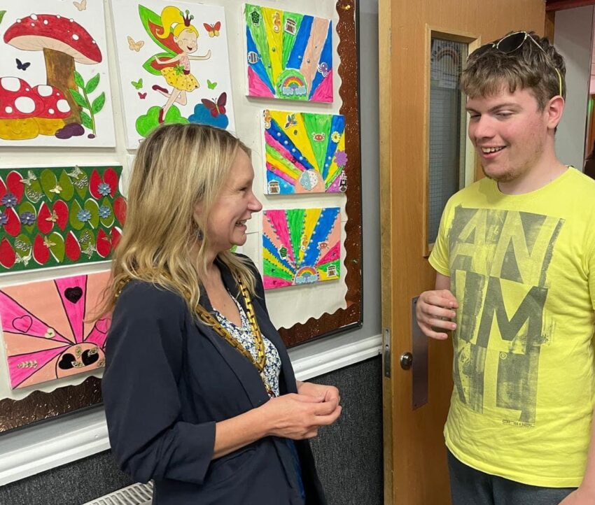 The Mayor, standing in front of a wall covered in colourful paintings, is talking to a young man in a yellow T-shirt